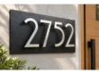 Black slate address sign with floating stainless steel 2752 numbers mounted on a light stucco wall beside a wooden door.