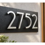 Black slate address sign with floating stainless steel 2752 numbers mounted on a light stucco wall beside a wooden door.