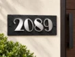 Black slate address plaque with raised silver stainless steel 2089 numbers beside a wooden entrance door.