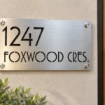 Brushed metal address sign with cut-out 1247 and FOXWOOD CRES. lettering on a stucco wall beside an oak door.