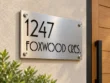 Left angled view of a brushed metal 1247 FOXWOOD CRES. sign showing cut-out lettering and round corner fixings.