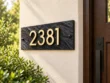 Right-angle view of a dark stone sign with gold-coloured 2381 numbers on a cream wall beside greenery.