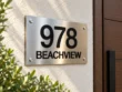 Left angled view of a brushed stainless steel 978 BEACHVIEW address plaque with raised black characters and silver fixings.