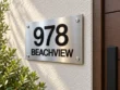 Right angled view of a horizontal stainless steel sign reading 978 BEACHVIEW on a light stucco wall beside a wooden door.