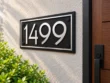 Right angled view of a black stainless steel address sign with silver 1499 numbers and border on a light stucco wall.