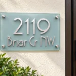 Modern Glass Address Sign - Frosted glass address sign with raised silver 2119 and Briar Cr NW lettering beside a wooden front door.