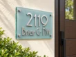 Right-angle view of the glass 2119 Briar Cr NW address sign mounted on a cream wall beside greenery.