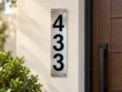 Slim stainless steel house number sign displaying 433 vertically on a cream wall beside a wooden entrance door.