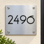 Square brushed stainless steel house number sign with black 2496 numerals on ivory stucco beside a wooden door.