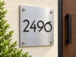 Left angled view of a square stainless steel 2496 address plaque showing black numeral edges and silver corner fixings.