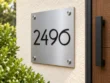 Right angled view of a square brushed stainless steel plaque with black 2496 numbers beside a wooden entrance door.