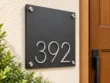 Left angled view of a matte charcoal address plaque showing raised silver 392 numbers and round corner fixings.