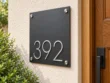 Right angled view of a square charcoal 392 address sign with raised silver numbers beside a wooden front door.