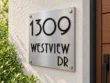 Right angled view of a square brushed stainless steel sign with black 1309 WESTVIEW DR lettering on a stucco wall.
