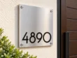 Right-angle view of a square brushed stainless steel 4890 address sign on a cream wall beside greenery and a wooden door.