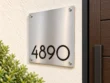 Left-angle view of a square stainless steel plaque with low-set black 4890 numbers and four silver corner fixings.