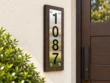 Left-angle view of a vertical 1087 stainless steel address plaque showing the dark wood backing and silver fixings.