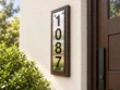 Right-angle view of a stainless steel 1087 house number sign on dark wood backing beside a front door and greenery.
