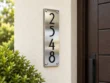 Right-angle view of a vertical stainless steel 2548 house number sign on a cream wall beside greenery.