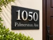 Left-angle view of a black slate 1050 Palmerston Ave sign showing floating silver numbers and textured stone.