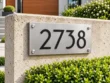 Left-angle view of a brushed stainless steel address plaque with black numbers 2738 and silver corner fixings.