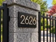 Left-angle view of a black 2626 address plaque with raised metallic numbers beside a residential gate.