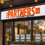 The Partners real estate light box sign with orange dimensional lettering and black signature text above a brick storefront entrance.