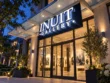 White illuminated Inuit Gallery channel letters mounted on a navy backing plate above a glass entrance at night.