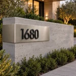Bent brushed stainless steel corner address sign 1680 with raised black numbers on a low concrete entrance wall