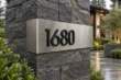 Bent stainless steel corner address sign 1680 with raised black numbers on a dark stone pillar