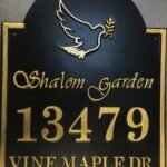 Arched black address sign reading Shalom Garden, 13479 Vine Maple Dr, with gold-coloured lettering, border and a dove carrying an olive branch.
