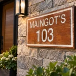 Wood-grain address sign reading MAINGOT’S 103 with white lettering and border, shown on a stone wall beside a front door and lantern.