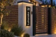 Angled view of a black aluminium 20 Luhanska address panel with illuminated cut-through characters beside a wood slat gate.