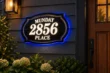 Classic black address plaque reading Munday 2856 Place, with white illuminated lettering, gold-tone border and blue halo on dark siding.