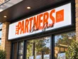 Wider storefront view of The Partners illuminated sign above glass doors and real estate listing displays.