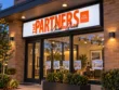 Angled close-up of The Partners orange dimensional lettering and black signature against a white illuminated sign face.