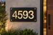 Black aluminium address plaque with warm light shining through cut-out numbers 4593, mounted on a stone wall with four silver-tone fixings.