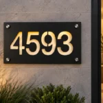 Black aluminium address plaque with warm light shining through cut-out numbers 4593, mounted on a stone wall with four silver-tone fixings.