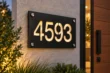 Angled view of a black aluminium 4593 address sign with warm illuminated cut-out numbers and silver-tone corner fixings beside a wooden door.
